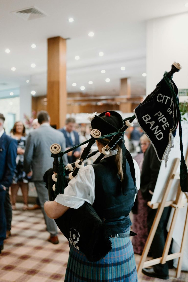 A bagpiper dressed in traditional attire playing at an indoor event with attendees in the background.