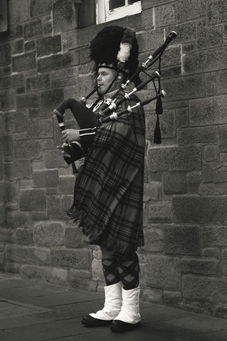 A Scottish bagpiper in traditional attire playing the pipes against a stone wall.